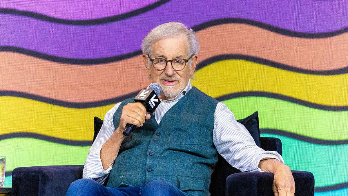 steven spielberg in a chair talking in a mic in front of rainbow background